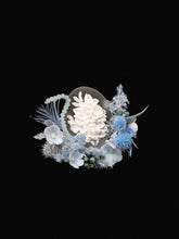 The Winteersweet Birth Flower Ring by CAMELLIA features a dried preserved flower arrangement with white blooms at the center, surrounded by blue and light purple petals against lush foliage on a black background.